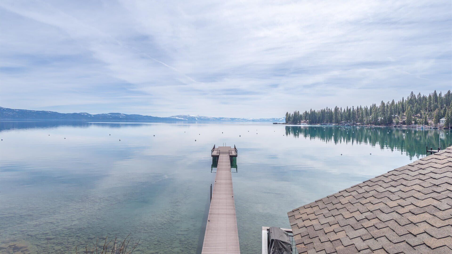 A clear view of a lake with a wooden dock extending into the water, surrounded by trees and distant mountains.