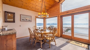 A wooden interior with a dining table and chairs, a chandelier, a framed picture, and a large window with a view of a lake and mountains.