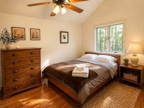A bedroom with a bed, wooden dresser, ceiling fan, framed pictures, window, and bedside lamp.
