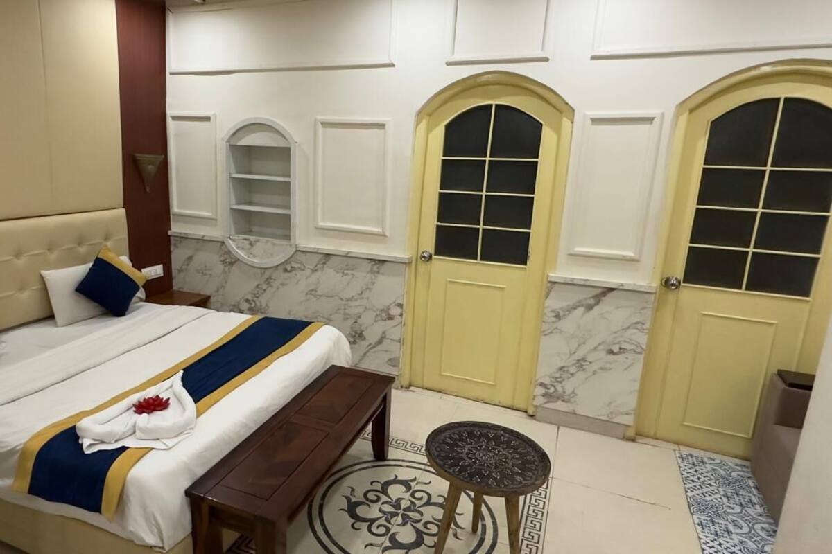 A hotel room with a bed, bedside table, bench, two doors, and a small round table.