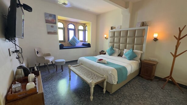 Habitación de hotel con cama, escritorio con silla, una mesita, un banco, perchero y ventana con vistas.