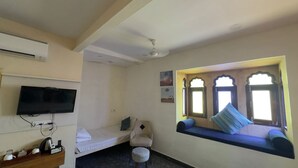 Una habitación con una cama, televisor, una mesita con hervidor y un rincón con ventanal y cojines.