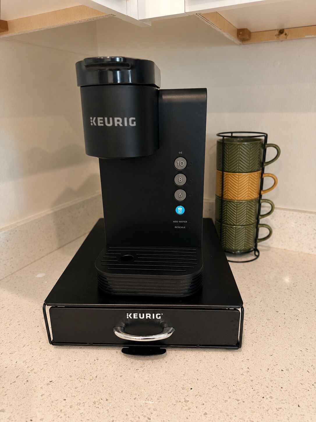 Une machine à café Keurig, montée sur un socle noir, avec un réservoir d’eau, un compartiment pour les dosettes de café et une tasse à café.