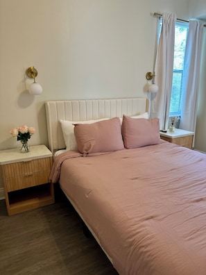 Un dormitorio con cabecera blanca, cama con ropa de cama rosa, mesita de noche con un florero y ventana con cortinas.
