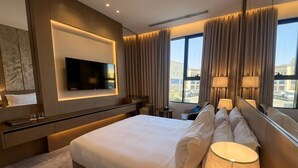 A modern hotel room with a large bed, a flat-screen TV mounted on the wall, a desk, and a view of the city through large windows. - La Terrasse Hotel (Jeddah)
