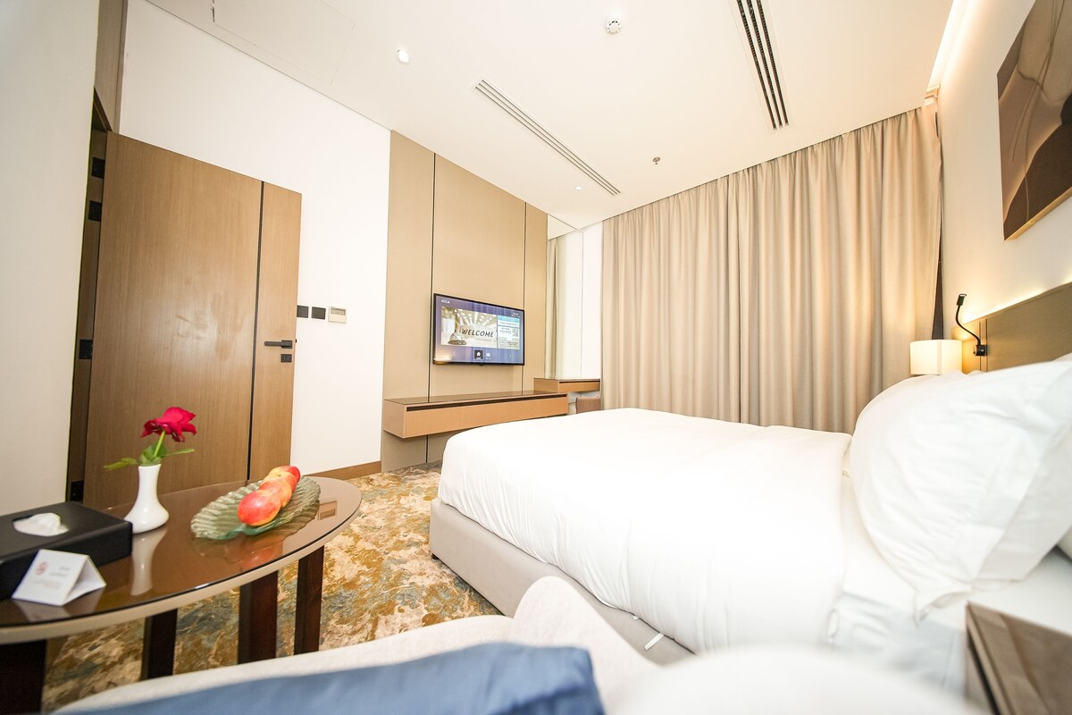 A hotel room with a bed, a TV, a desk with a vase and fruit, and a door.