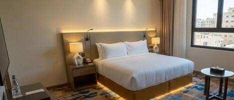 Habitación de hotel con una cama grande, lámparas de noche, una mesita, un escritorio y vistas a la ciudad a través de la ventana.