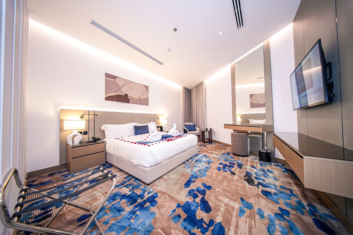 A modern hotel room with a large bed, bedside tables, a desk, a chair, a TV, and a patterned carpet.