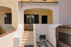 Eine Treppe führt zu einem überdachten Außenbereich mit Tisch und Stühlen, einem Fenster mit Rollläden und einer Tür.