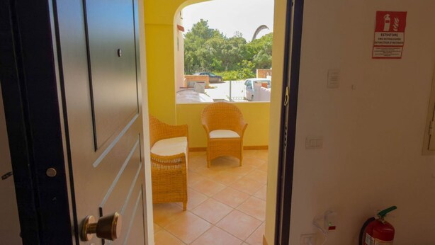 Une pièce avec une porte donnant sur un balcon équipé de chaises en osier et d’un extincteur fixé au mur.