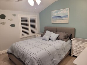 Une chambre à coucher comprenant un lit, des tables de chevet, un ventilateur de plafond, une fenêtre avec des stores et des éléments de décoration murale.