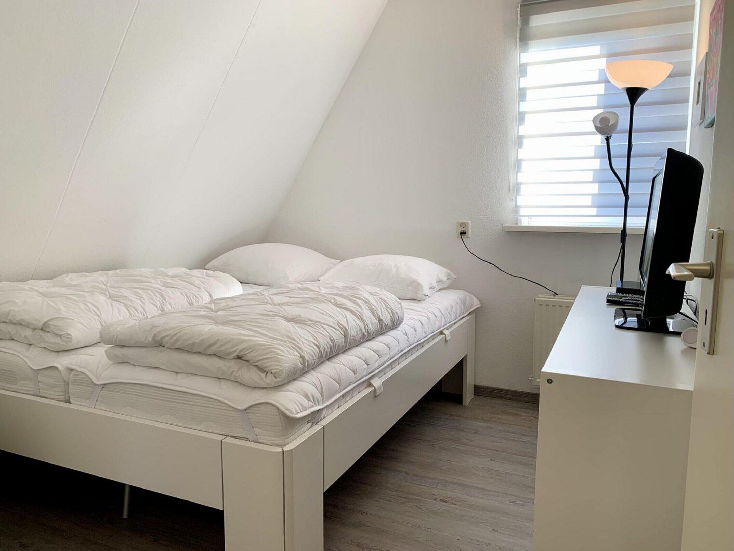 A white, single-bed room with a sloped ceiling, a desk with a computer, and a window with blinds.