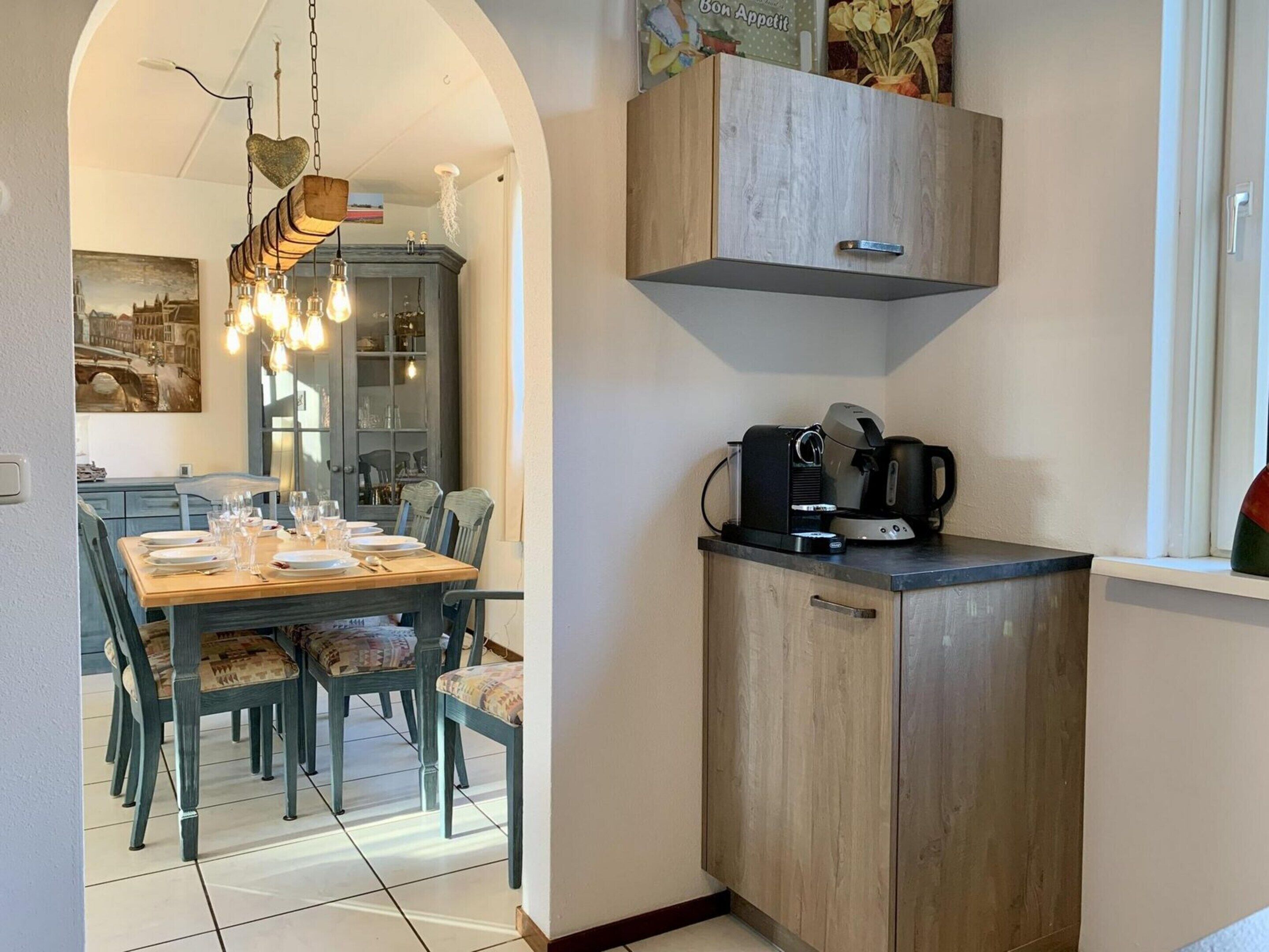 A dining area with a wooden table and chairs, a kitchen with a coffee machine, and a cabinet above the counter.