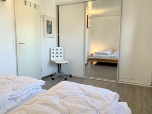 Quarto de hotel com duas camas, uma cadeira branca, um espelho grande e piso de madeira.