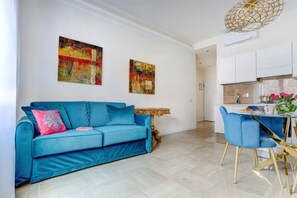 Ein modernes Wohnzimmer mit einem blauen Sofa, zwei abstrakten Gemälden, einem Essbereich mit einem weißen Tisch und blauen Stühlen sowie einer Küche im Hintergrund.