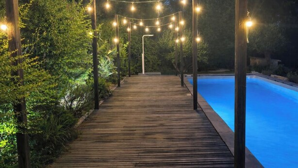 Un camino de madera que conduce a una piscina, rodeada de follaje y luces colgantes.