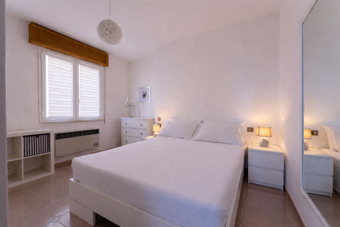 Un dormitorio con una cama grande, mesitas de noche, cómoda, una ventana con contraventanas, una lámpara de techo e imagen colgada en la pared.