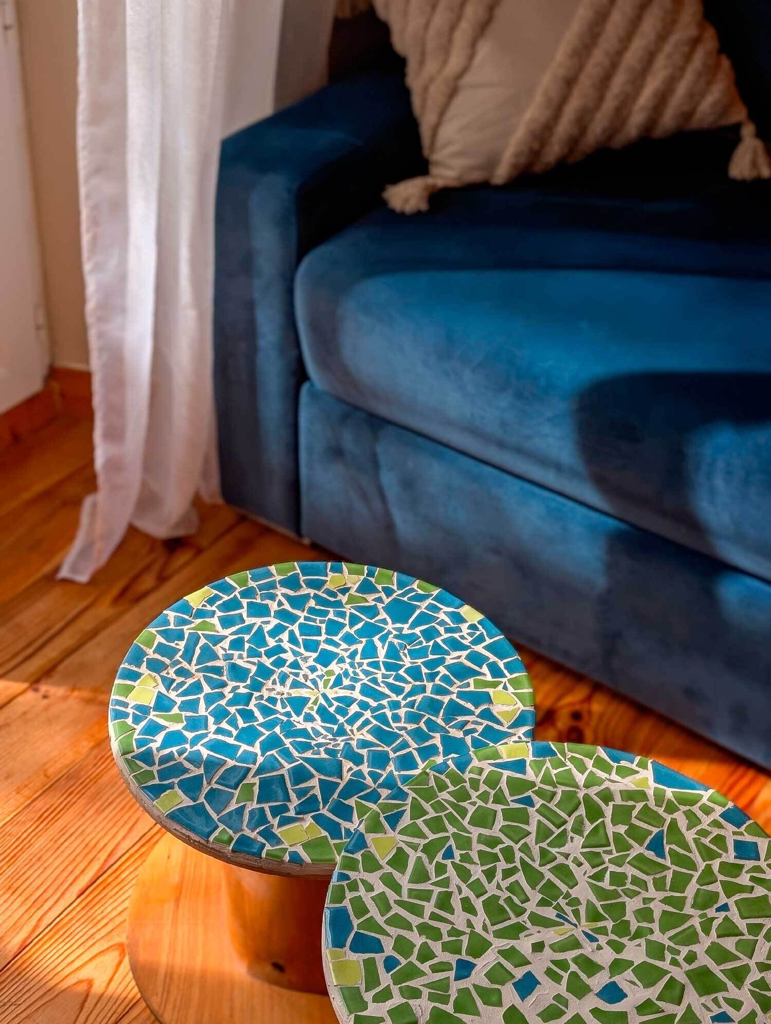 A blue sofa with a white cushion, a white curtain, and two mosaic-tiled round tables on a wooden floor.