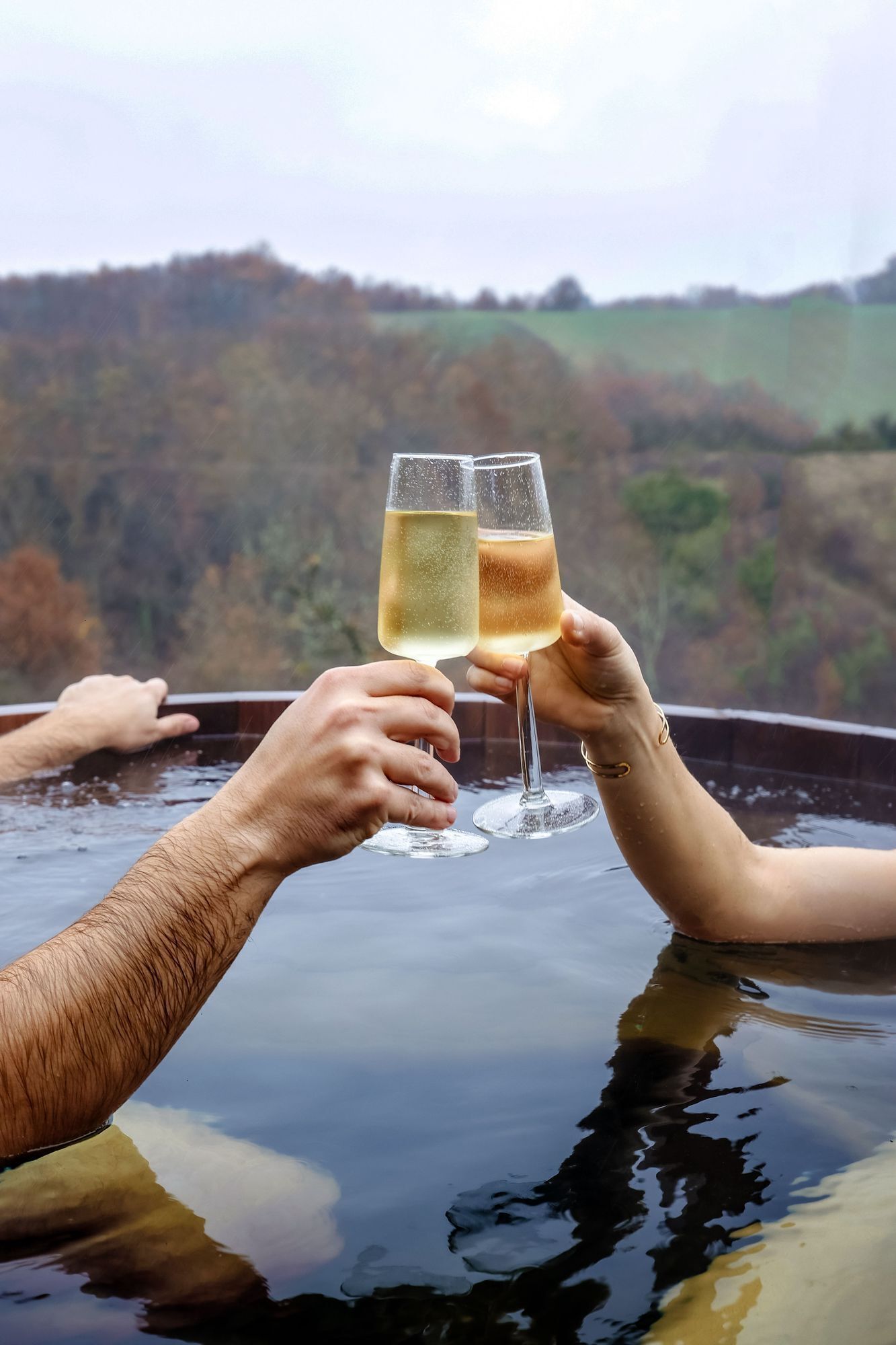Two people toasting with champagne glasses in a hot tub with a scenic view of hills.