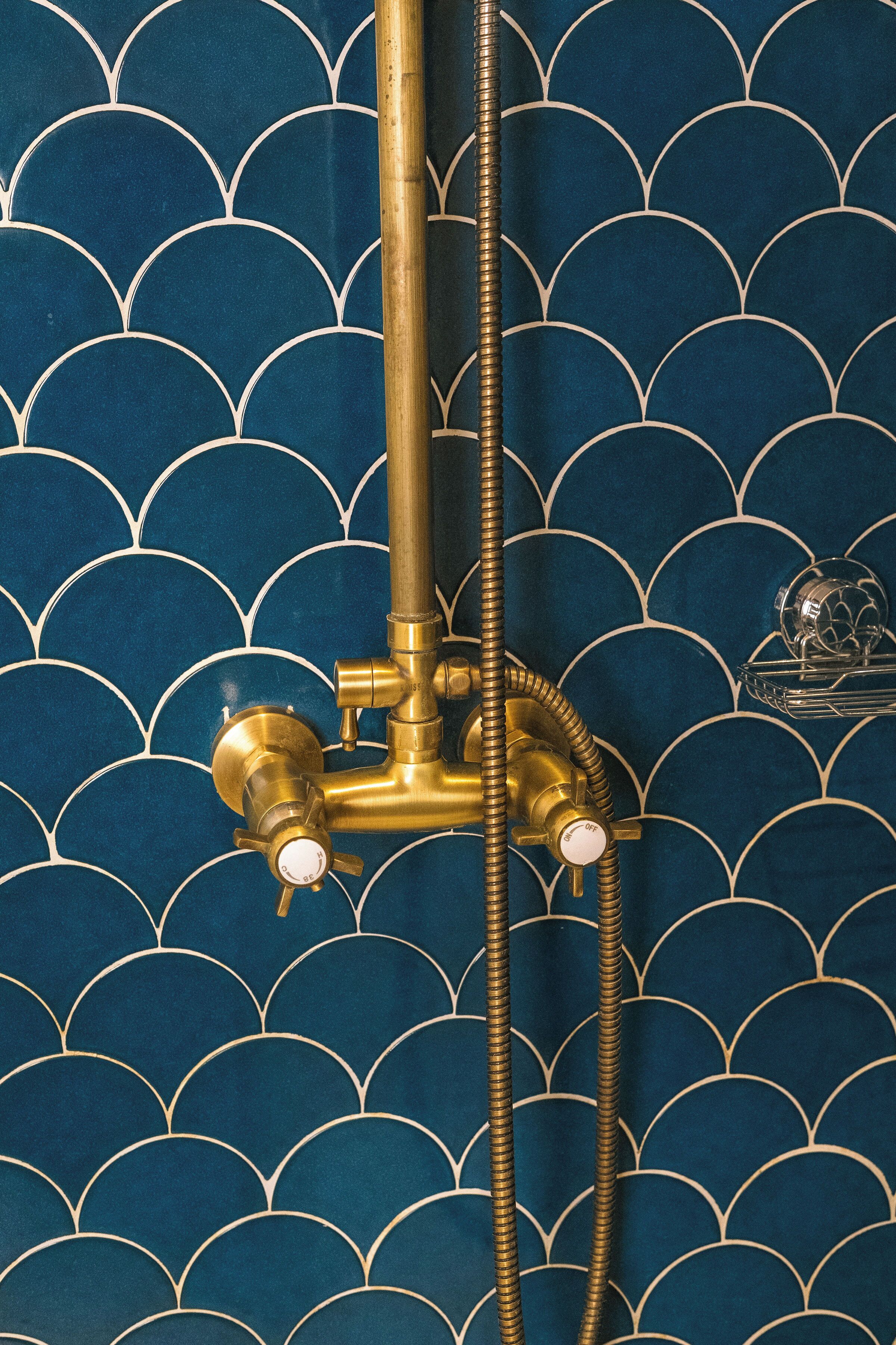 Ein Badezimmer mit blauen Fliesen an der Wand, die ein geschwungenes Muster aufweisen, einer goldfarbenen Duschbrause und einem Handduschteil.
