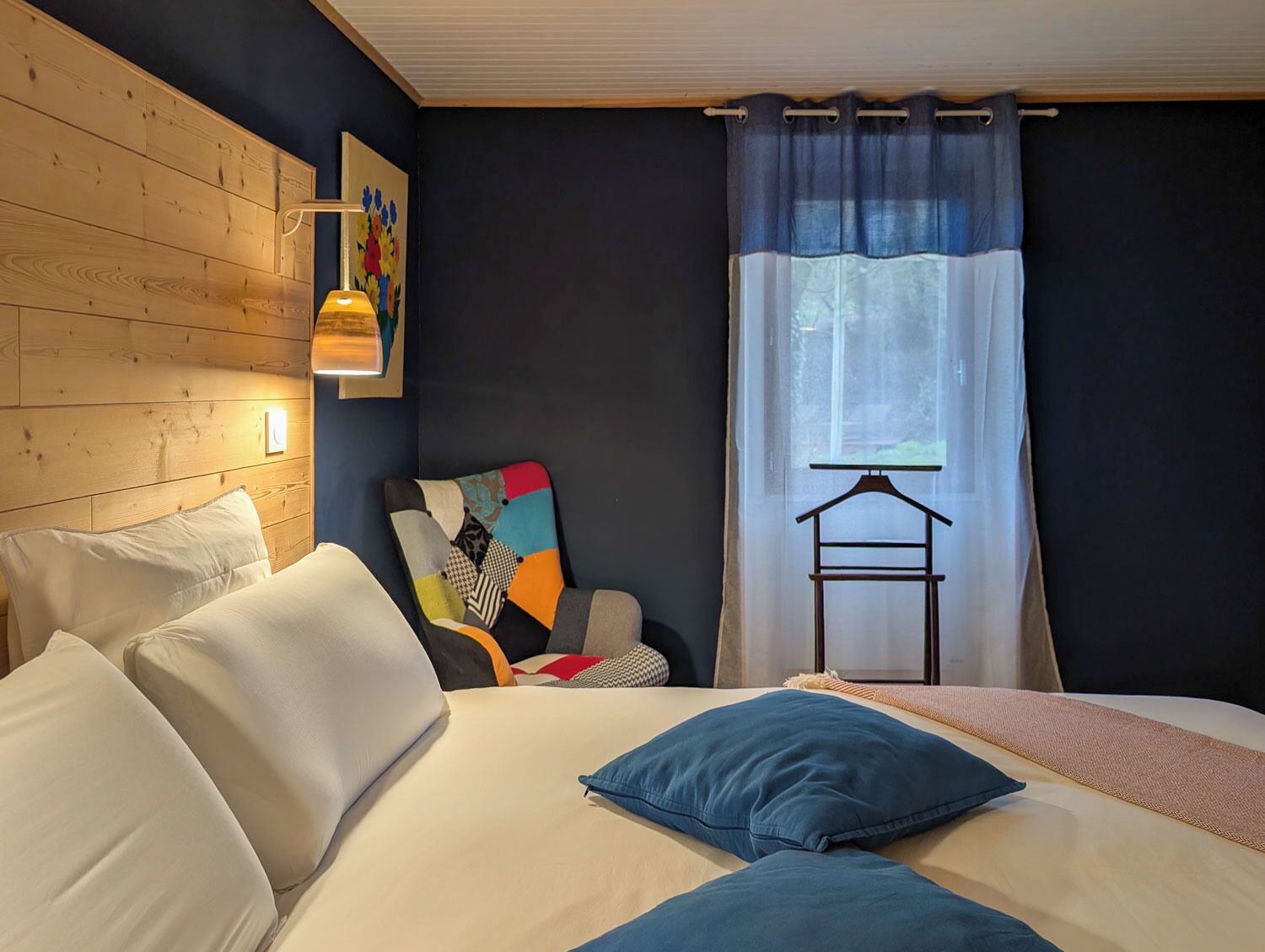 A bedroom with a bed, pillows, a chair with a colorful cushion, a hanging light, and a window with curtains.