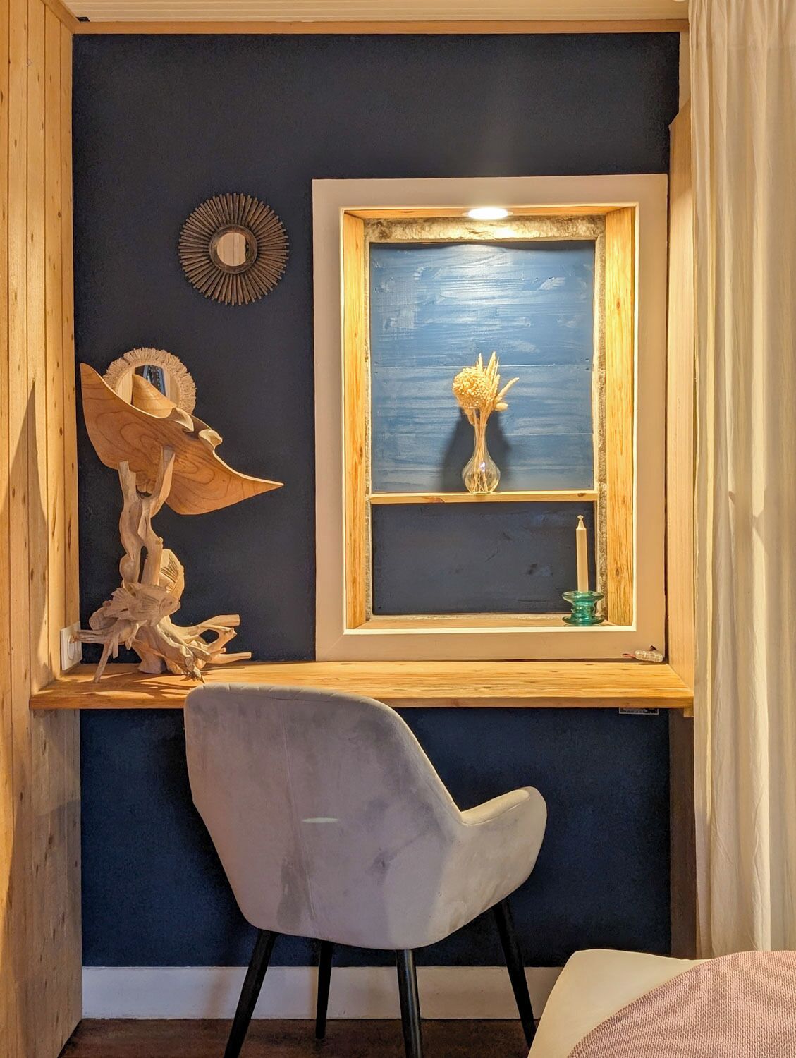 A room with a dark blue wall, a wooden desk with a chair, a window with a view, and decorative items.
