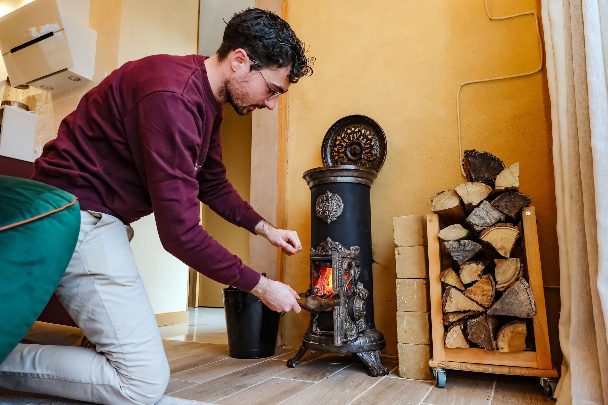 Eine Person bedient einen Holzofen in einem Raum mit Fliesenboden und einem Holzstapel.