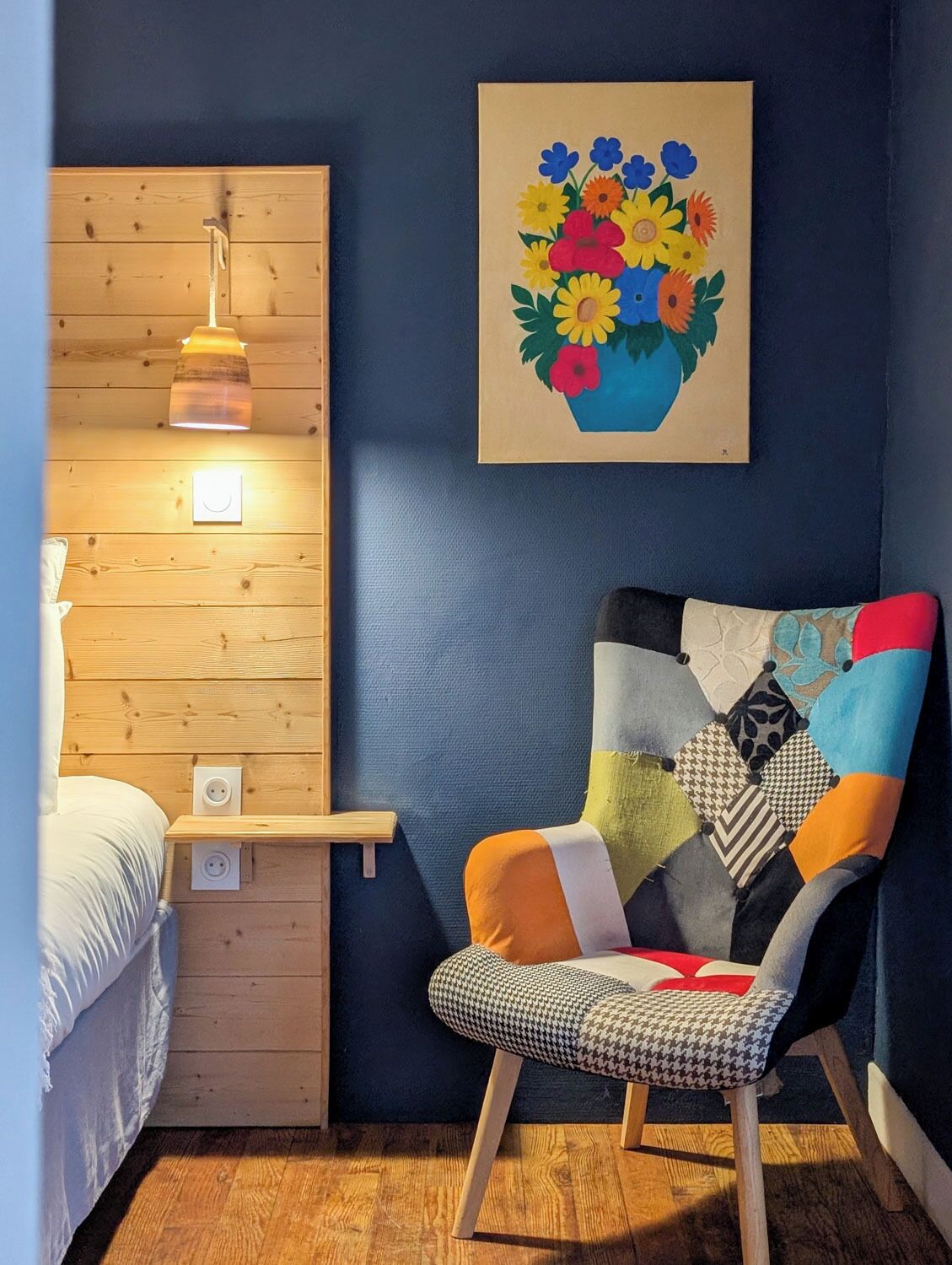 Una stanza con una parete in legno, una sedia con un cuscino patchwork, un quadro di fiori e una lampada da comodino.