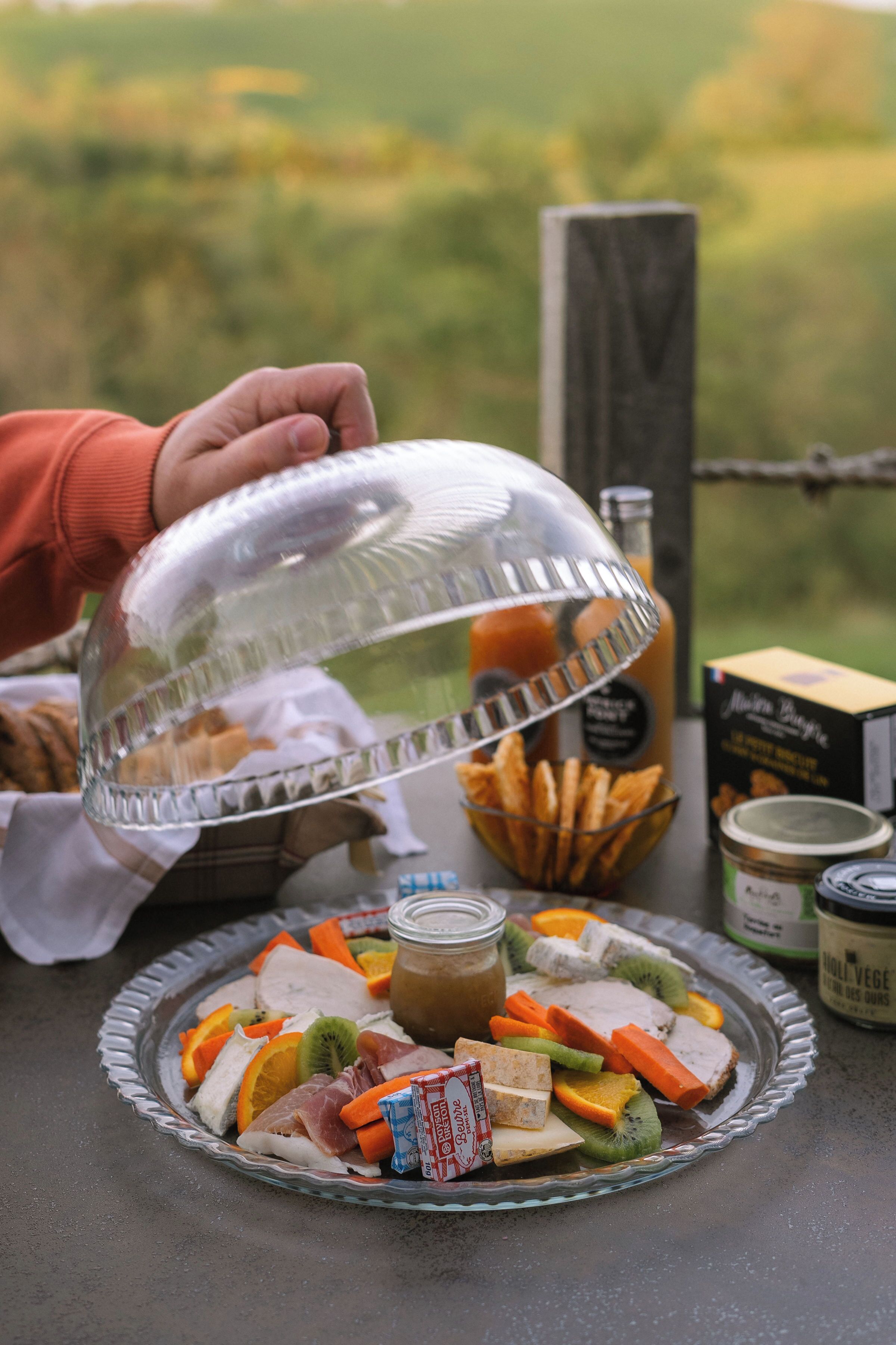 A glass pie dish with assorted fruits and a jar of jam on a table with a clear glass dome.