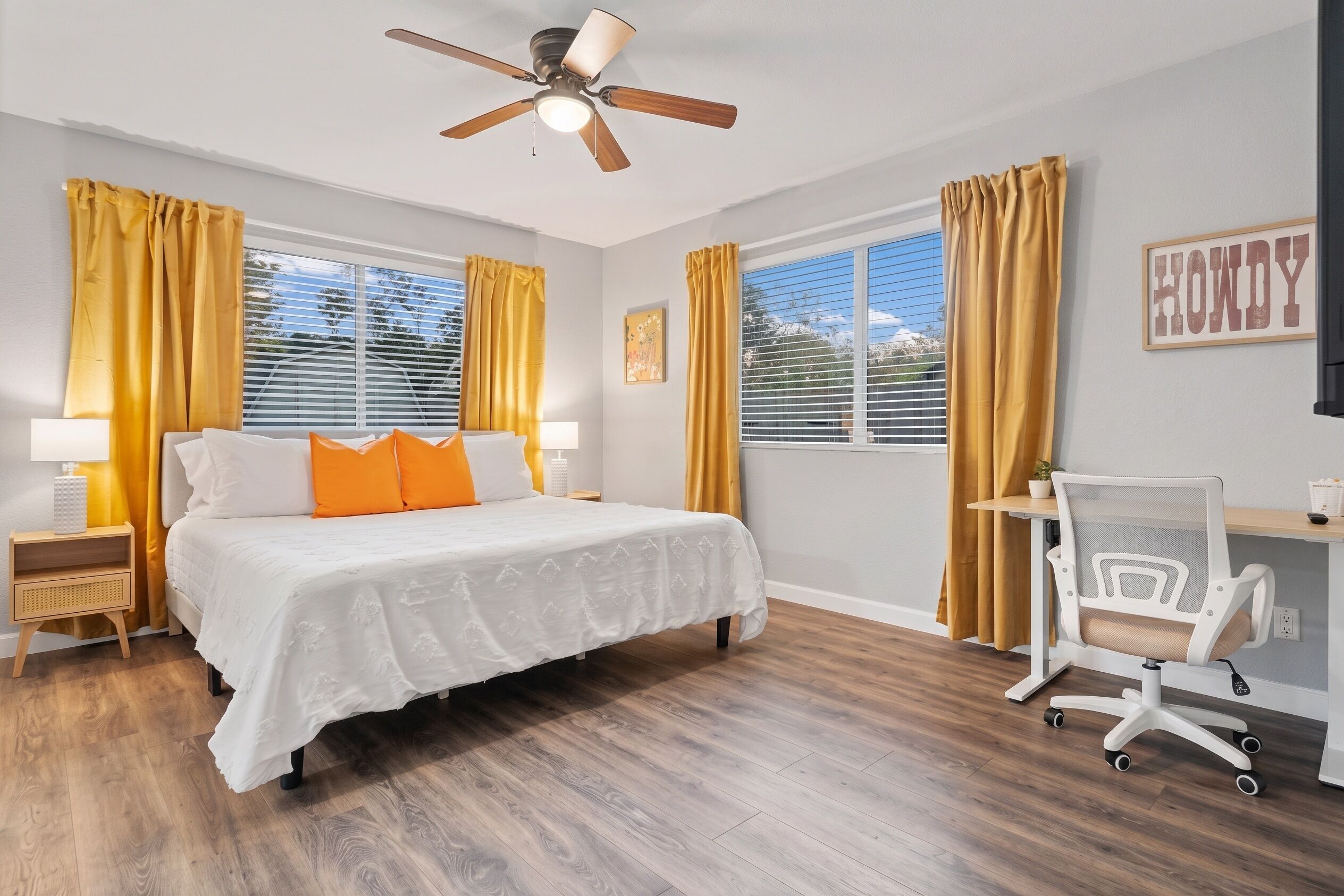 Une chambre à coucher comprenant un lit, un bureau, une chaise, un ventilateur de plafond et des rideaux.
