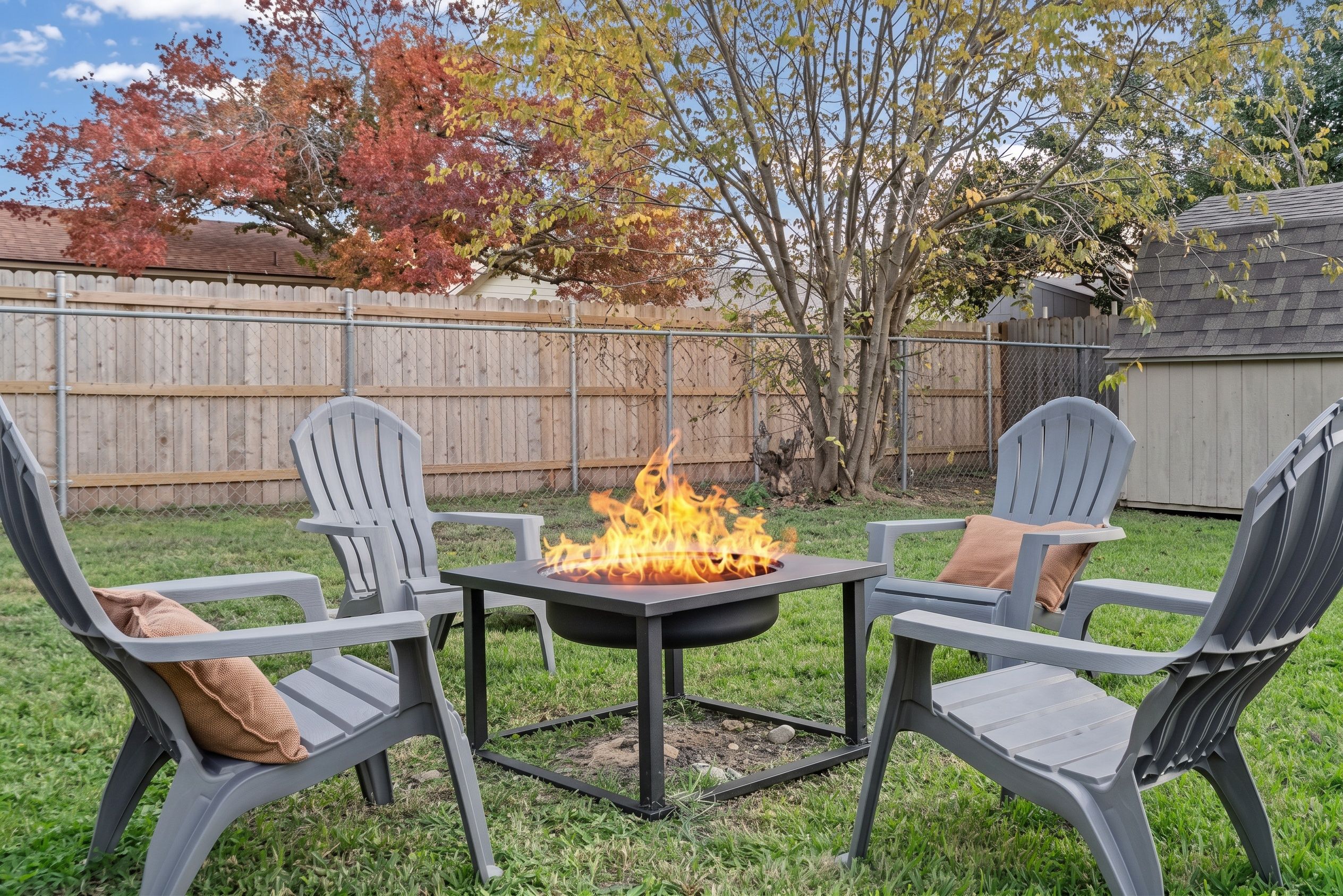 A backyard with a fire pit table surrounded by Adirondack chairs with cushions.