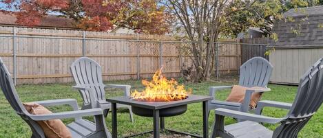 A backyard with a fire pit table surrounded by Adirondack chairs with cushions.