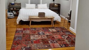 Una camera da letto con un letto, una ventola a soffitto, una finestra con persiane, un comodino, una sedia, un tappeto e una libreria.