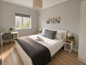 Une chambre à coucher comprenant un lit, des tables de chevet, un bureau, une chaise, une fenêtre et des œuvres d’art accrochées au mur.