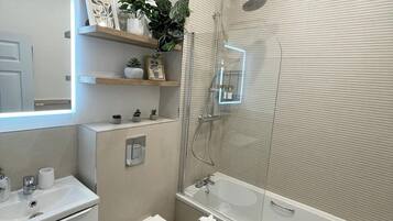 A modern bathroom with a white sink, toilet, and bathtub. There are shelves with plants and decorative items, and a shower with a glass enclosure.