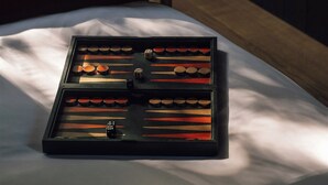 Un lit bien fait, avec un jeu de société posé au centre, un oreiller rouge, une petite table sur laquelle se trouvent une bouteille et des verres, et une fenêtre avec des rideaux.