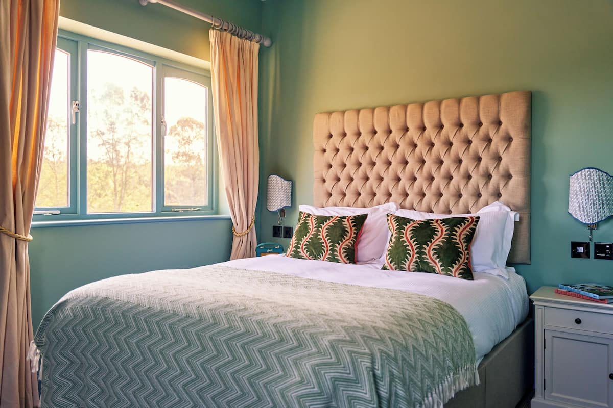 A bedroom with a large bed, a window with curtains, a nightstand with a lamp, and a bedside table with books.