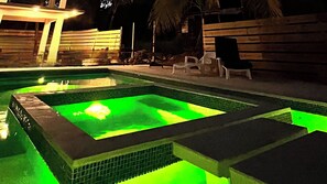 Una piscina con iluminación verde, rodeada de cubierta y sillas de madera.