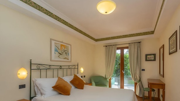Un dormitorio con cama, dos mesitas de noche, una silla verde, una pequeña mesa y una ventana con cortinas.