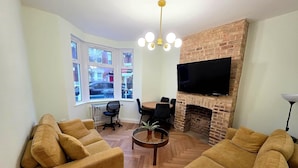 A living room with a fireplace, a TV mounted on the wall, and a dining area with chairs. - Modern 2BDR Home with Garden in Walthamstow (Greater London)