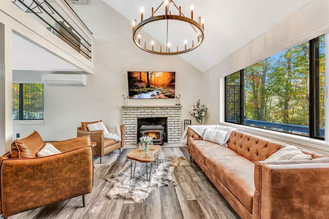 A modern living room with a fireplace, leather sofas, a chandelier, and a large window with a forest view.