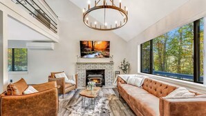 A modern living room with a fireplace, leather sofas, a chandelier, and a large window with a forest view. - Bushkill Bliss w/ Hot tub near Falls & Shawnee (East Stroudsburg)