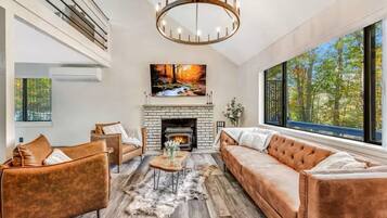 A modern living room with a fireplace, leather sofas, a chandelier, and a large window with a forest view.