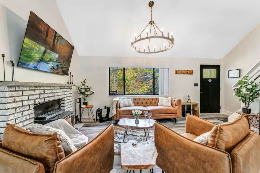 A living room with a fireplace, leather sofas, a chandelier, and a large window with a view of trees.