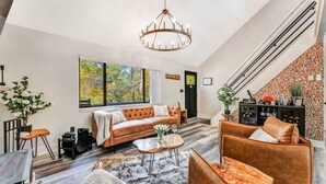 A spacious living room with a large window, a leather sofa, a coffee table, and a chandelier. - Bushkill Bliss w/ Hot tub near Falls & Shawnee (East Stroudsburg)