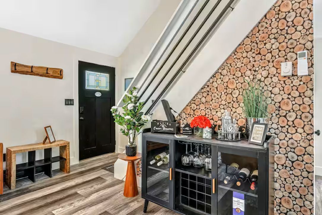 A modern interior with a staircase, a black door, a wooden bench, a console table with wine bottles, and a wall decorated with logs.