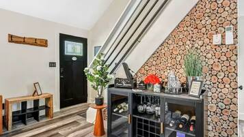 A modern interior with a staircase, a black door, a wooden bench, a console table with wine bottles, and a wall decorated with logs.