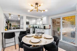 A modern kitchen with white cabinets, a dining area with a round table set for four, and a large sliding glass door leading to an outdoor patio. - Charming Cabin in the Woods w/Hot tub & Firepit (Albrightsville)