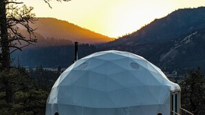Estructura de domo geodésico con una ventana circular central, rodeada por un paisaje boscoso y montañas al fondo.