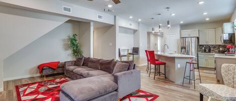 A modern living room with a sectional sofa, a red and white patterned rug, a ceiling fan, and a kitchen area with a refrigerator and bar stools.